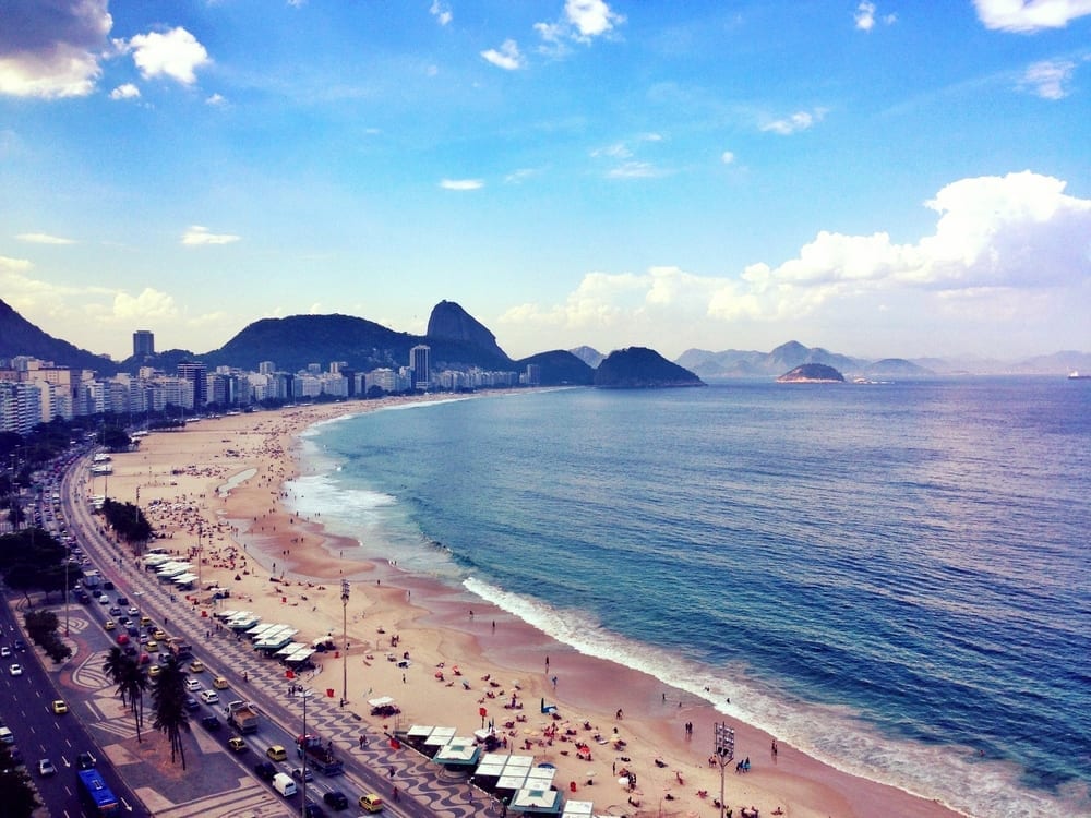 a beach with cars and buildings in the background with Copacabana, Rio de Janeiro in the background