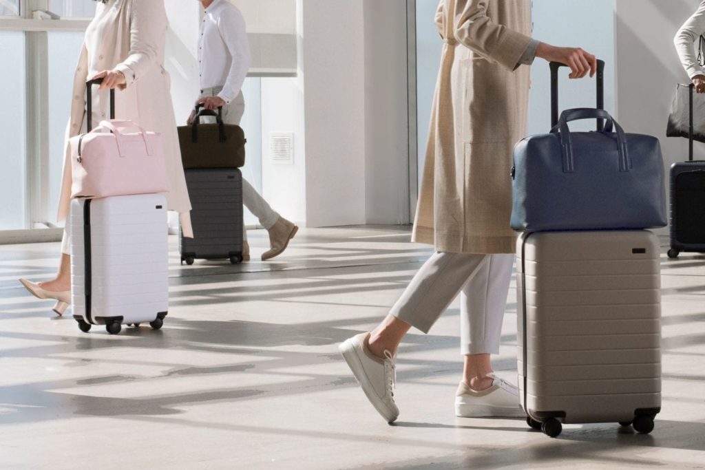 a woman in a beige coat and white pants walking with luggage