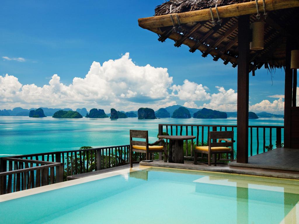 a pool with a thatched roof and chairs overlooking the ocean