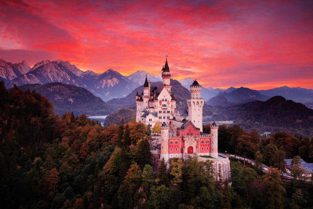 a castle on a hill with trees and mountains in the background with Neuschwanstein Castle in the background