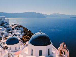a white buildings with blue domes and a body of water with Santorini in the background