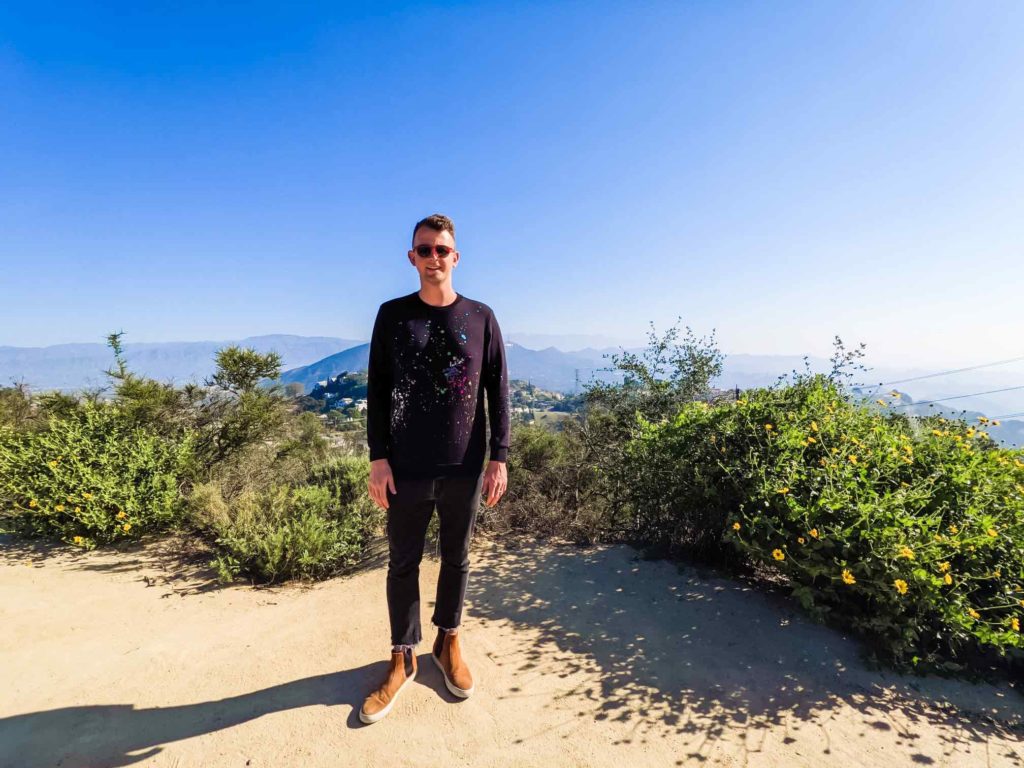 a man standing on a dirt path with bushes and mountains in the background