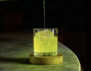 a glass with a yellow liquid and ice on a coaster